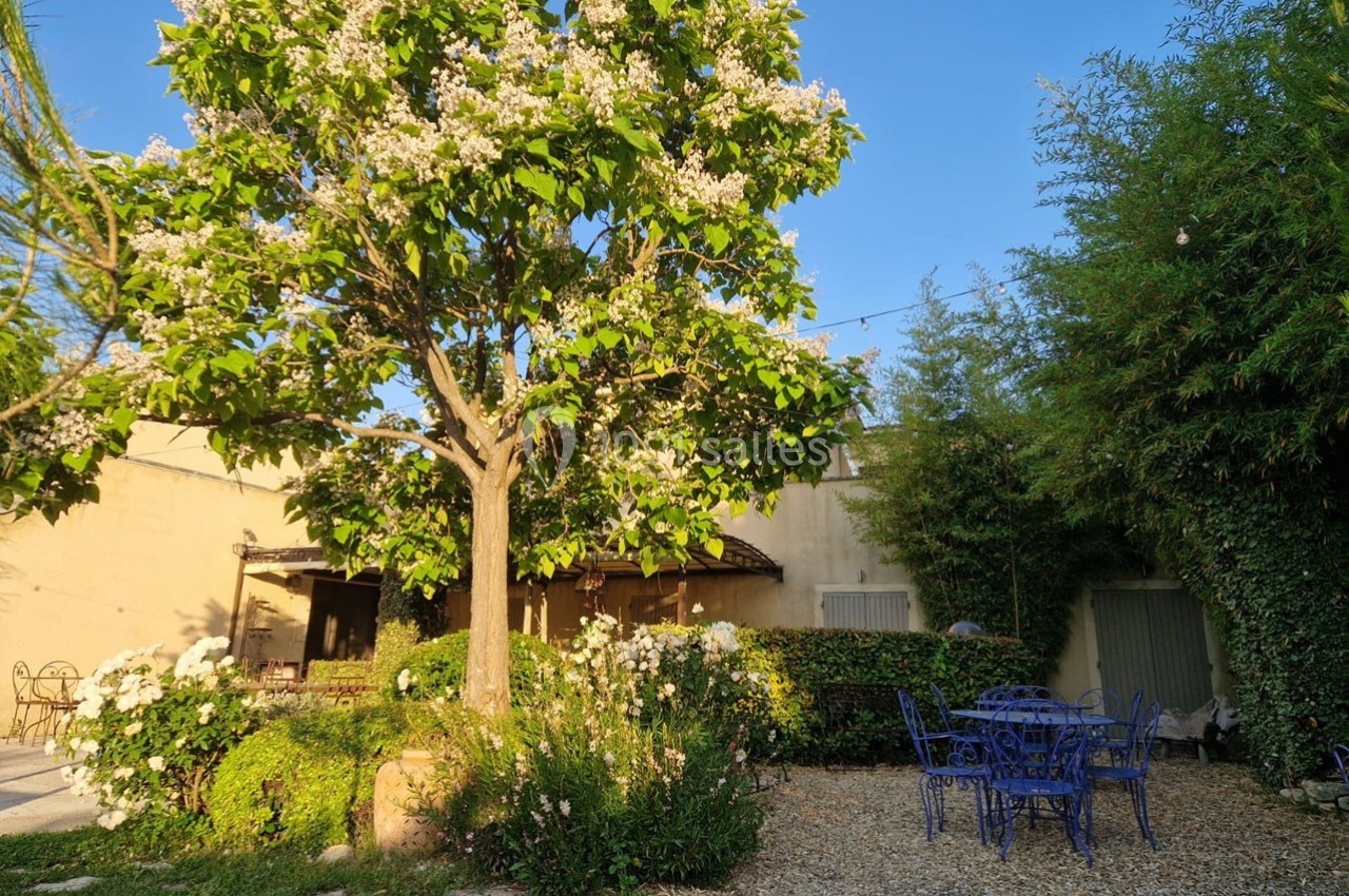 Jardin avec un arbre en fleurs, des buissons fleuris, une table et des chaises bleues sur un sol en gravier.