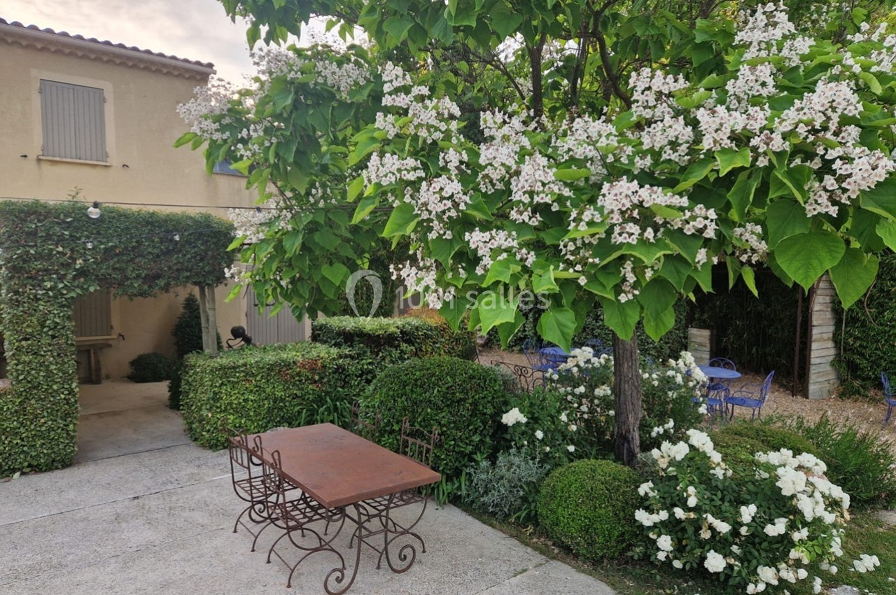 Cour extérieure avec table en fer forgé, chaises, arbre en fleurs et végétation soignée devant une maison beige.