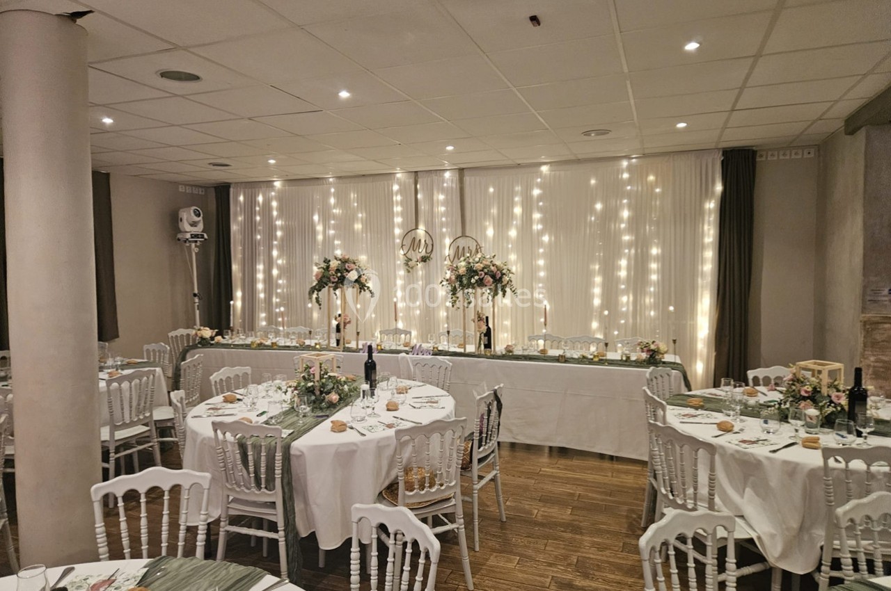 Salle de réception décorée pour un événement, avec tables rondes dressées et guirlandes lumineuses en arrière-plan.