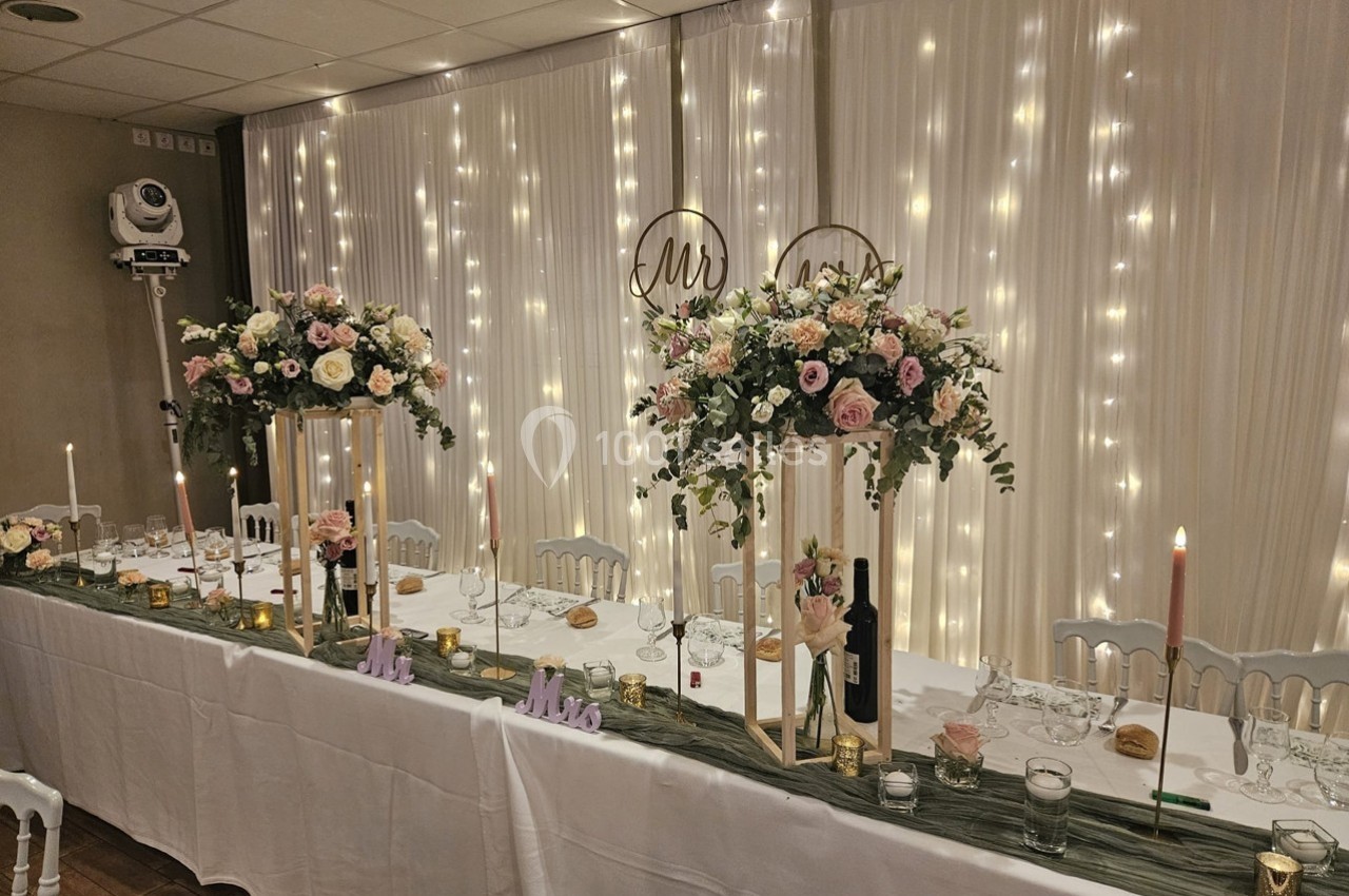Table de réception décorée avec des fleurs, bougies et guirlandes lumineuses, dans une salle élégante.