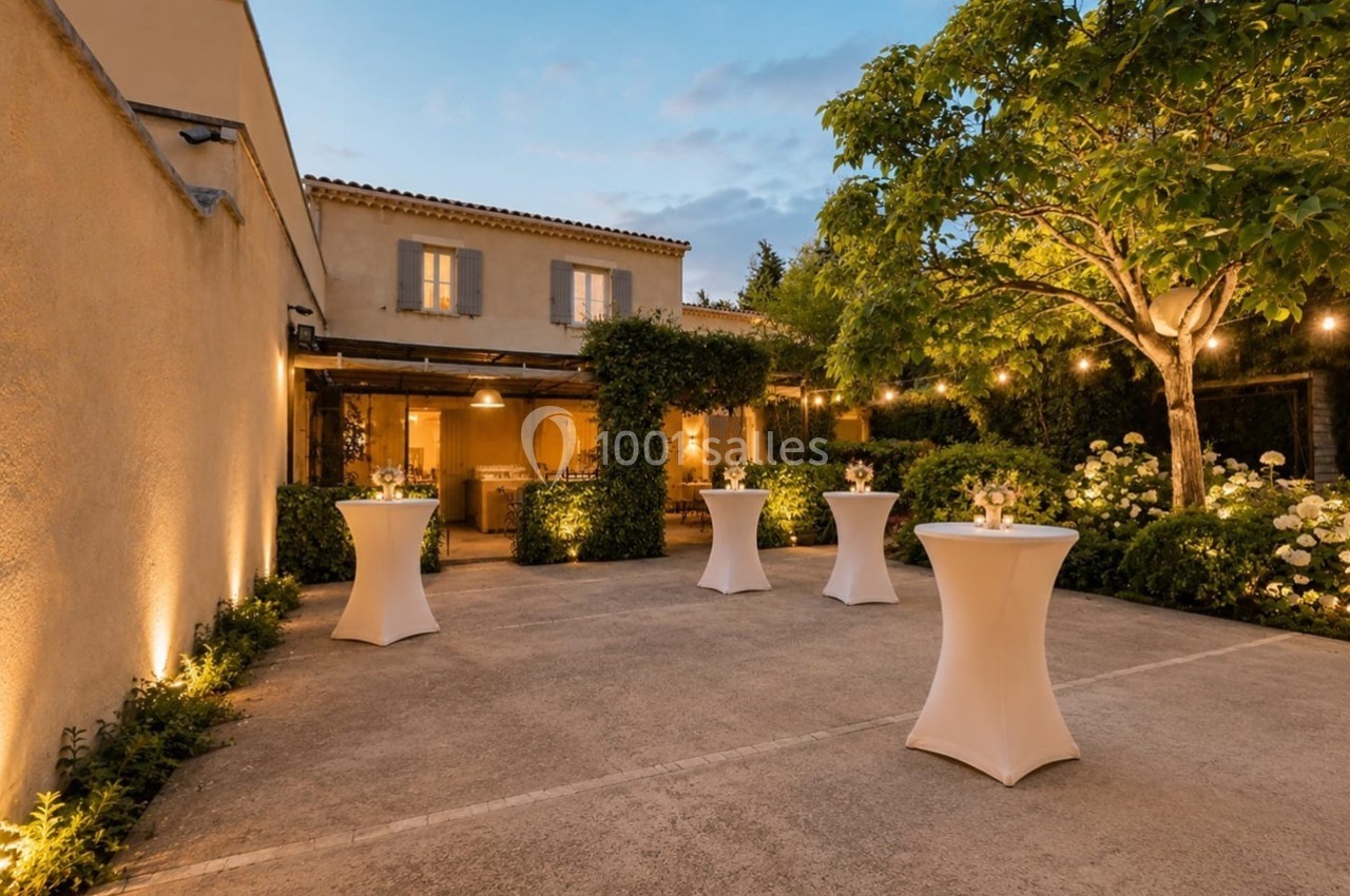 Cour extérieure d'une maison avec tables hautes décorées, éclairage chaleureux et végétation environnante.