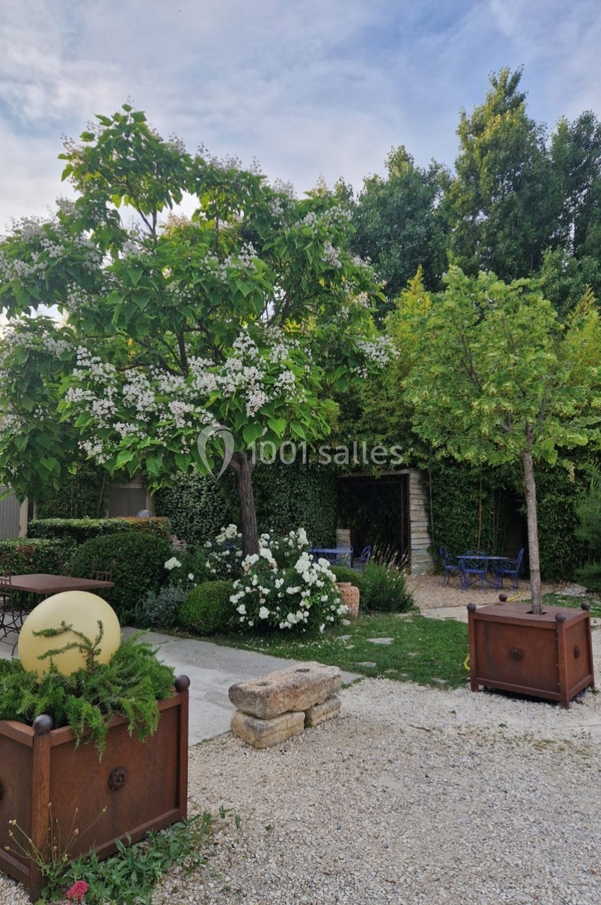 Jardin avec arbres en fleurs, buissons taillés, pots décoratifs et mobilier de jardin sur un sol gravillonné.
