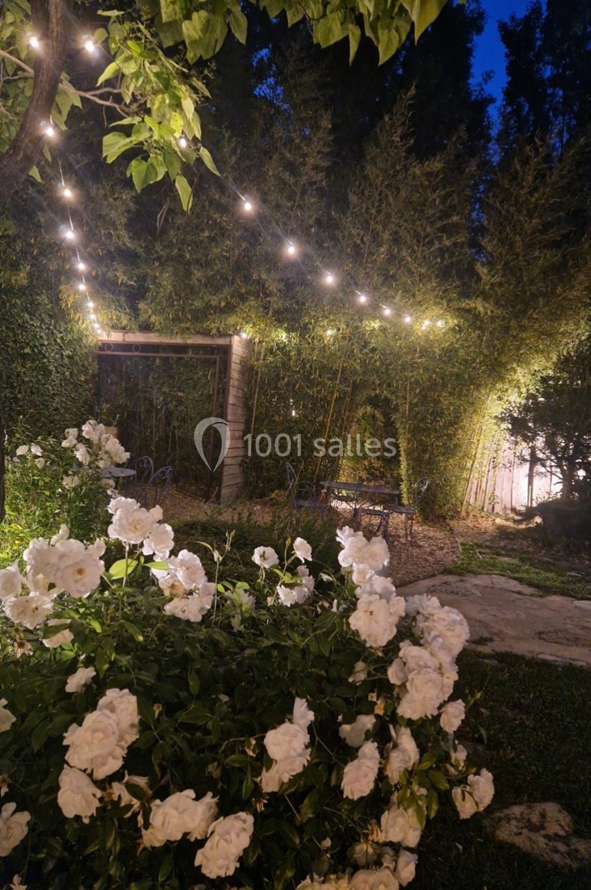 Guirlandes lumineuses suspendues dans un jardin fleuri avec des roses blanches et des arbres en arrière-plan.