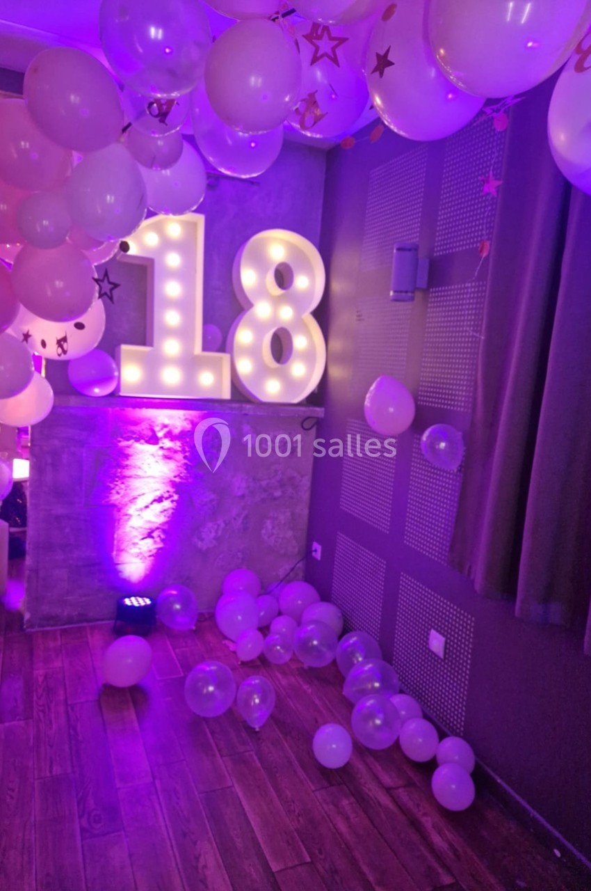 Décoration festive avec ballons roses et blancs, chiffres lumineux ’18’ et éclairage violet dans une pièce.
