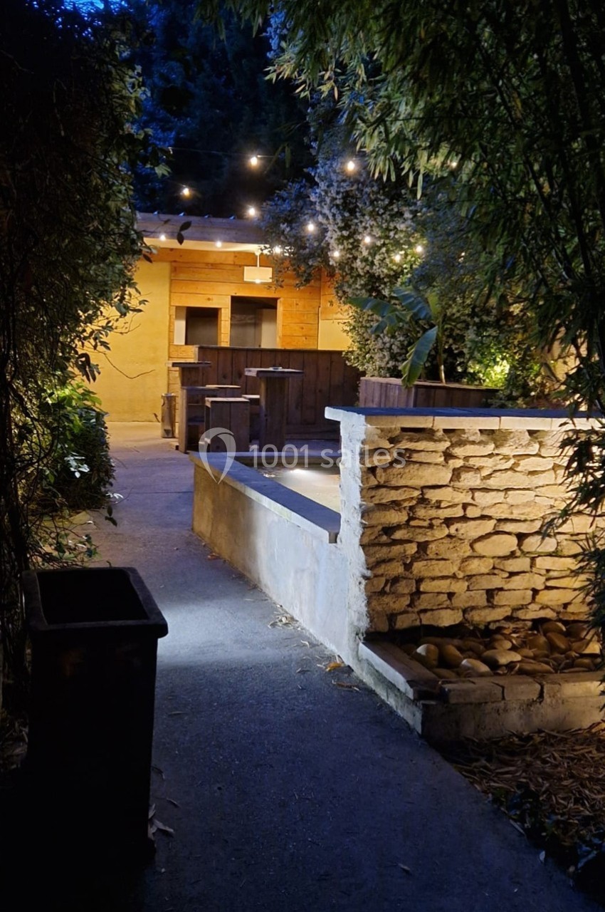 Allée éclairée menant à une terrasse en bois, entourée de végétation et décorée de guirlandes lumineuses.