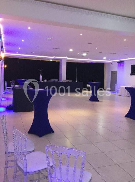 Salle de réception vide avec tables hautes drapées de bleu, chaises transparentes et éclairage tamisé.