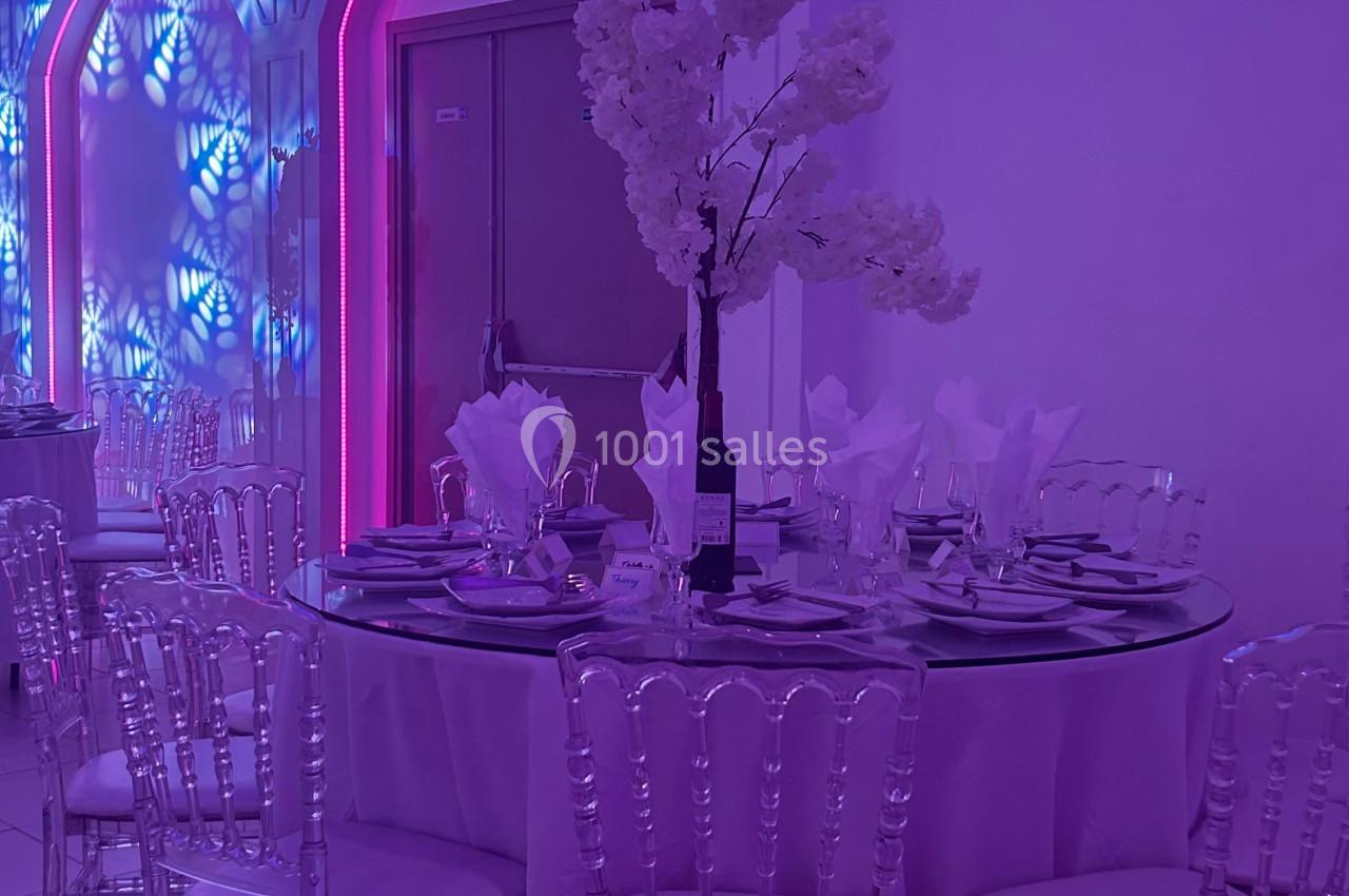 Salle de réception avec table ronde décorée, chaises transparentes et éclairage violet et bleu en arrière-plan.