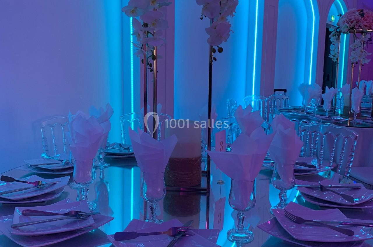 Table ronde décorée avec des assiettes, verres et serviettes, éclairée par une lumière bleue et ornée de fleurs.