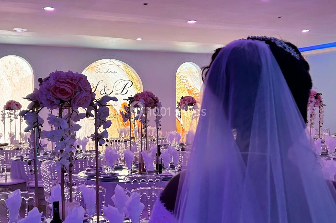 Une mariée de dos dans une salle de réception décorée avec des tables dressées et des arrangements floraux.