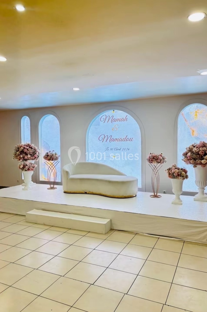 Décor de salle avec canapé blanc, fleurs roses sur piédestaux et inscription murale célébrant un mariage.