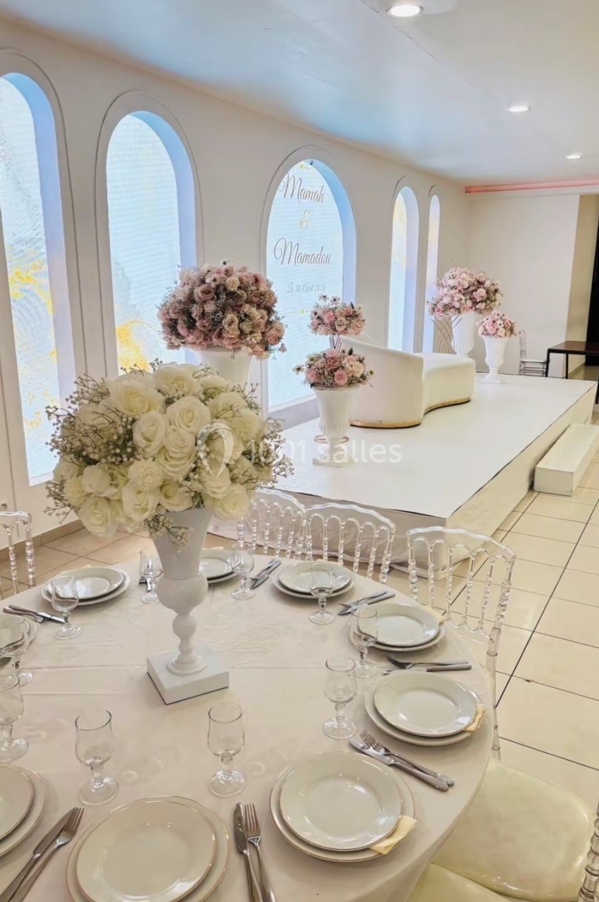 Salle de réception décorée avec des fleurs blanches et roses, table dressée et estrade avec canapé en arrière-plan.