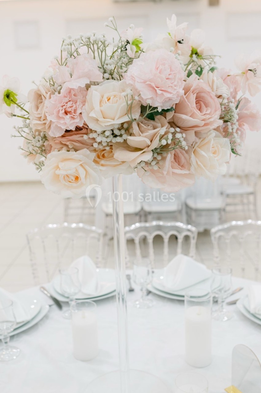 Centre de table floral avec roses pastel et gypsophile sur un support transparent, entouré de vaisselle blanche.