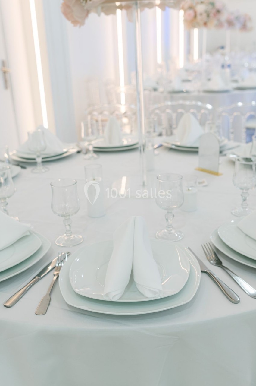 Table dressée avec nappes blanches, assiettes, couverts, verres et serviettes pliées dans un décor lumineux et élégant.
