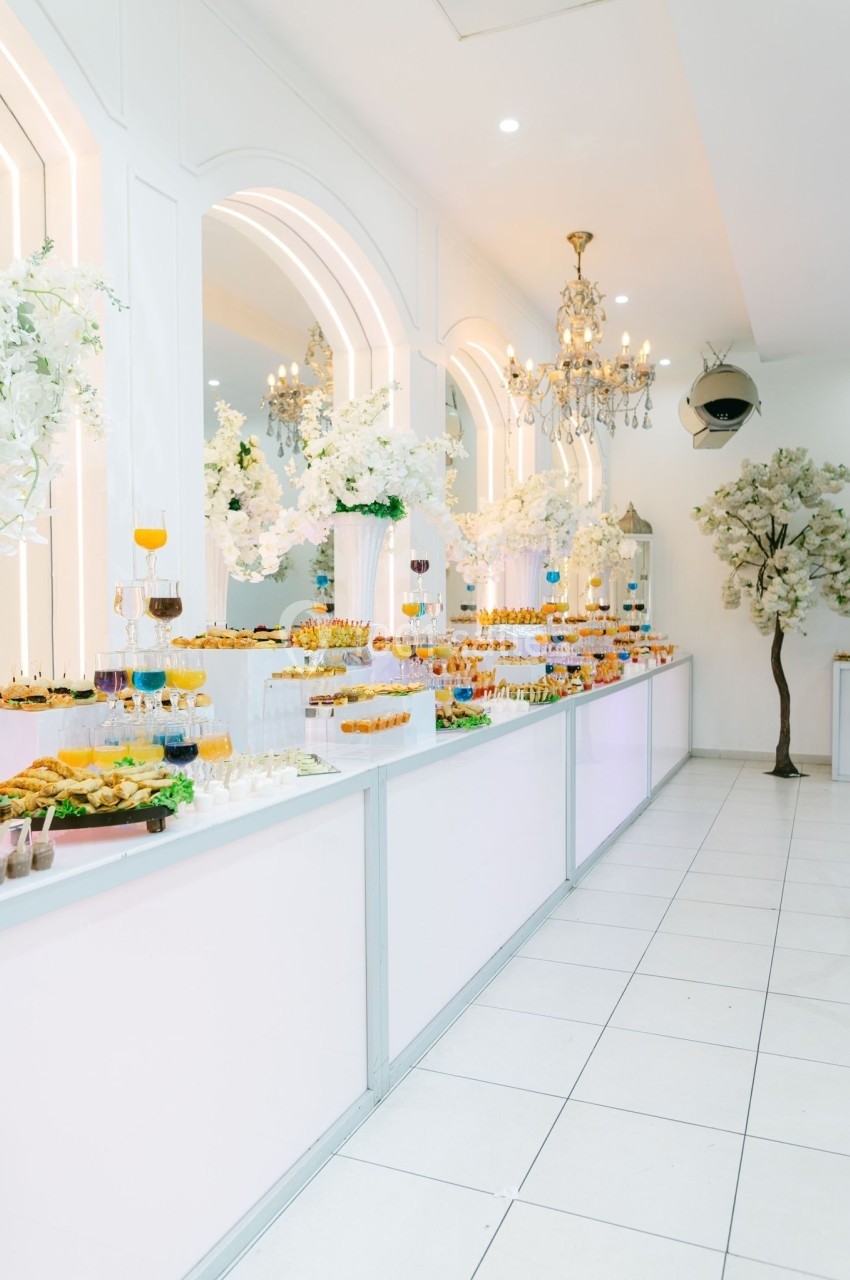 Salle lumineuse avec buffet de desserts variés, décorée de fleurs blanches, miroirs et lustres élégants.