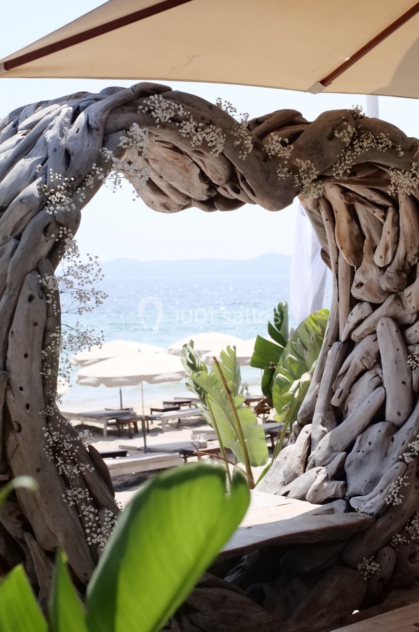 Structure en bois flotté ornée de fleurs blanches, encadrant une vue sur la mer avec parasols et végétation tropicale.