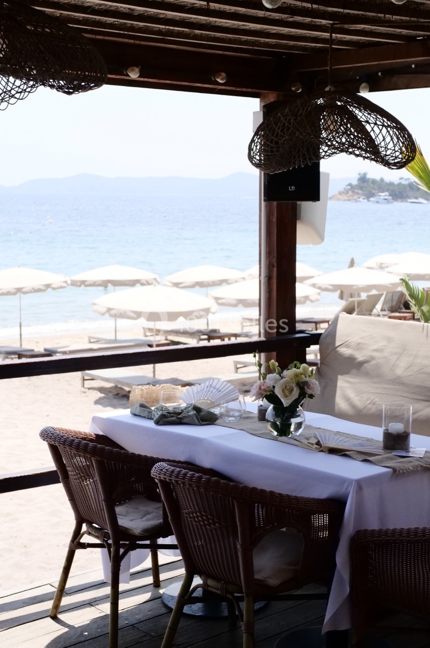 Table dressée sous une pergola en bois avec vue sur une plage, des parasols blancs et la mer en arrière-plan.