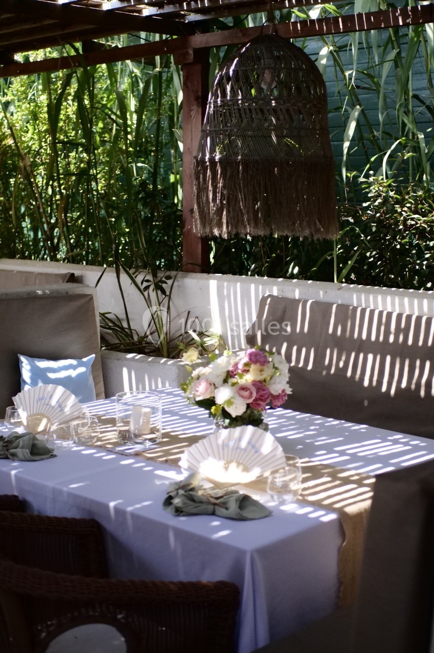 Table dressée en extérieur avec nappes blanches, bouquet de fleurs au centre et lumière filtrée par une pergola.