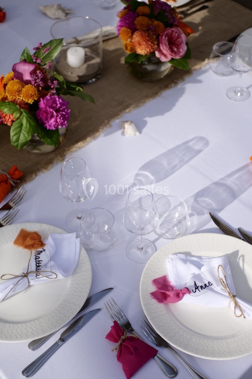 Table dressée avec nappes blanches, assiettes, couverts, serviettes personnalisées et décorations florales colorées.