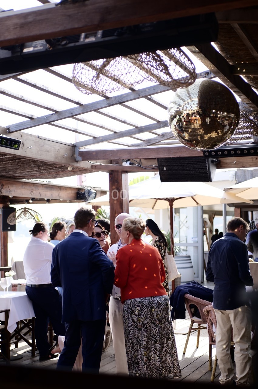 Personnes discutant sous une pergola en bois avec une boule disco suspendue et des tables dressées en arrière-plan.