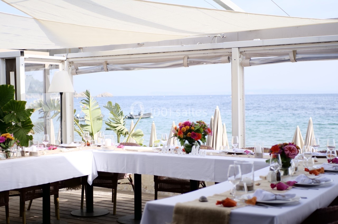 Salle de restaurant en bord de mer avec tables dressées, fleurs colorées et vue sur l'eau et le ciel dégagé.