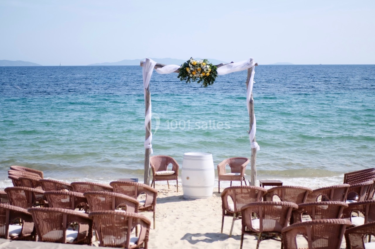 Arche décorée de fleurs face à la mer, entourée de chaises en osier disposées sur une plage de sable.