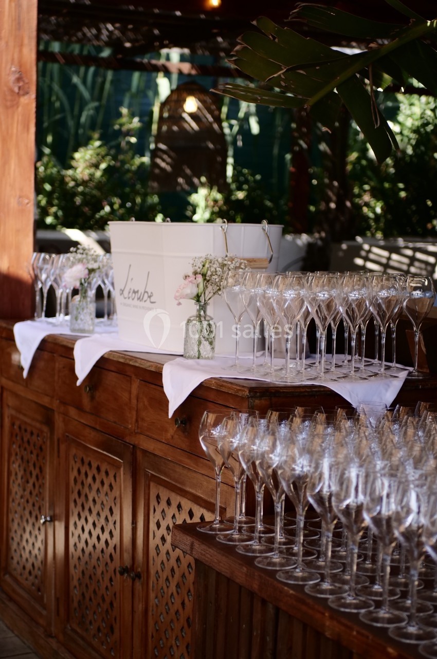 Table en bois avec seau à champagne, verres à pied alignés et décoration florale dans un cadre extérieur verdoyant.