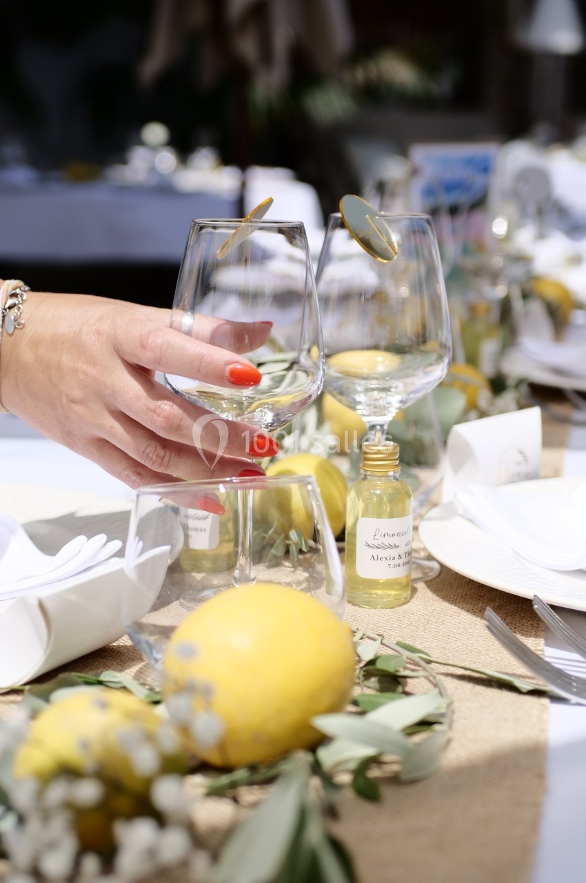 Main tenant un verre à pied sur une table décorée de citrons, feuillages et éléments de vaisselle.