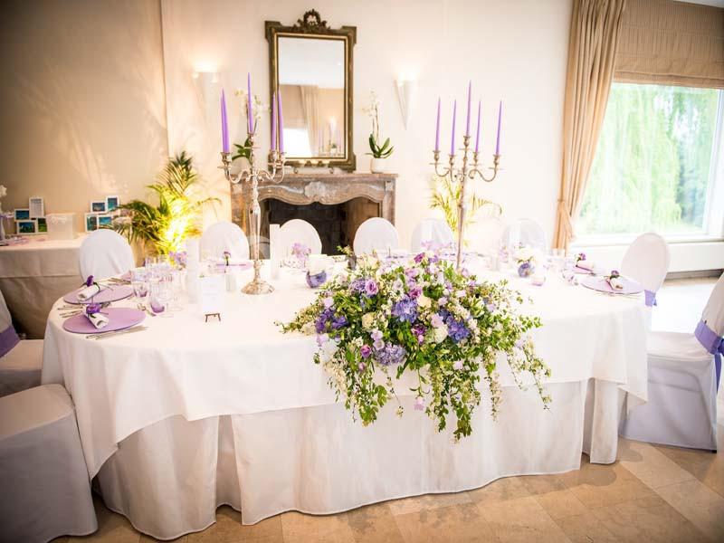 Table élégamment dressée avec nappes blanches, chandeliers, fleurs violettes et décoration raffinée dans une salle lumineuse.