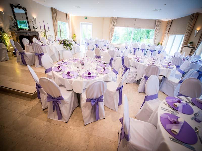 Salle de réception décorée avec des nappes blanches et des rubans violets, tables dressées pour un événement.