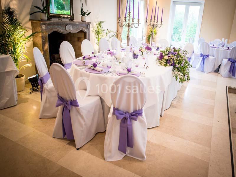 Salle de réception décorée avec des tables rondes, nappes blanches, chaises ornées de nœuds violets et chandeliers.