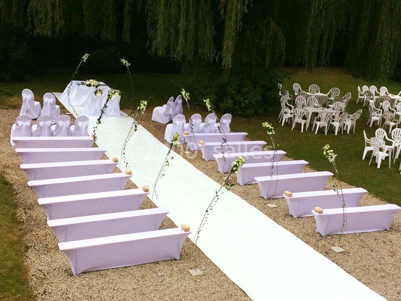 Allée centrale blanche entourée de bancs décorés pour une cérémonie en plein air, avec chaises disposées en arrière-plan.