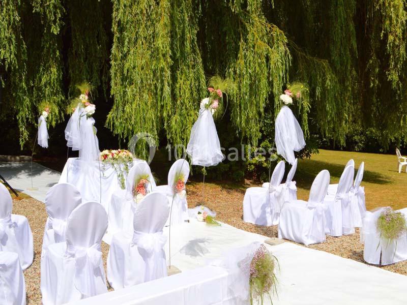 Chaises blanches décorées et alignées en extérieur sous des arbres, préparées pour une cérémonie ou un événement.
