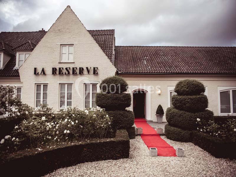 Façade d'un bâtiment blanc avec toit en tuiles, jardin soigné et tapis rouge menant à l'entrée principale.