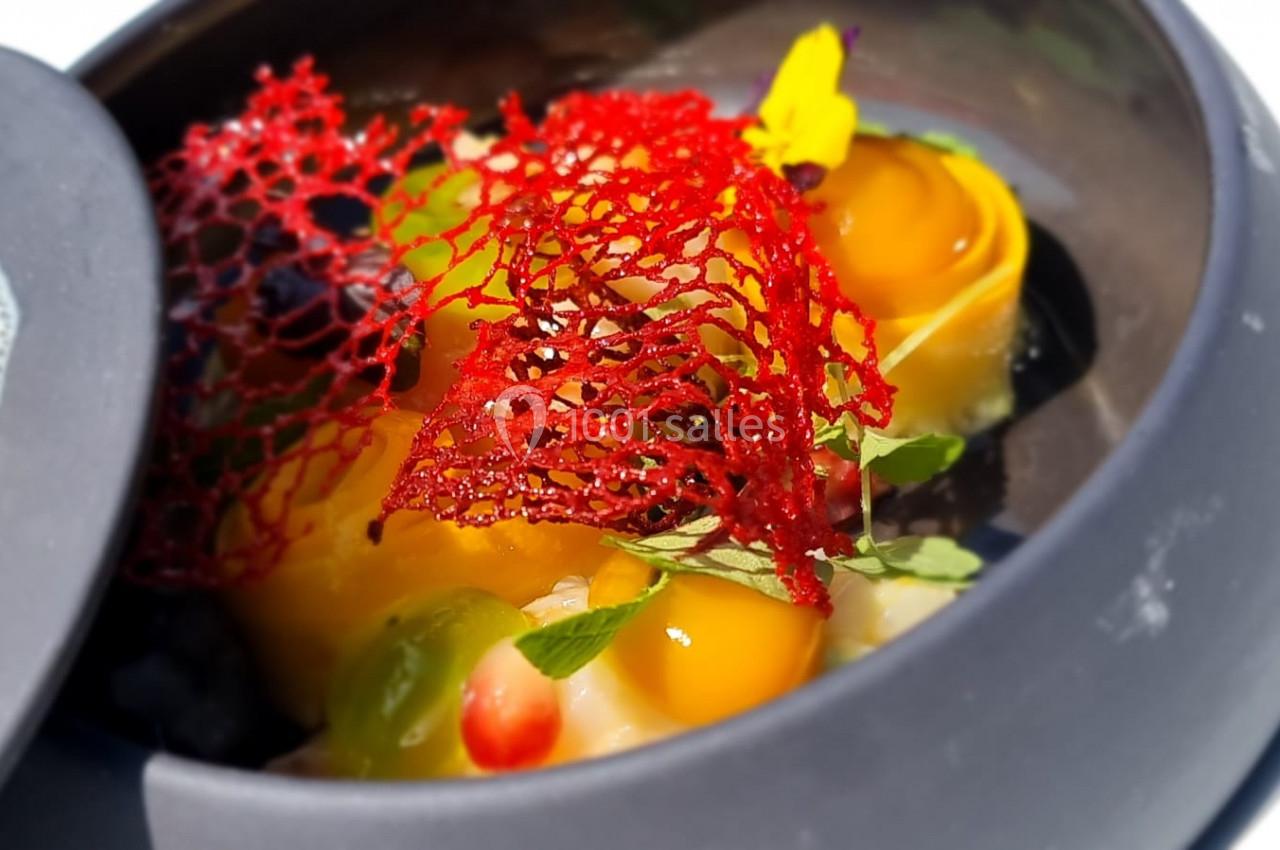 Plat gastronomique coloré dans une assiette noire, avec mousse, fleurs comestibles et décor en dentelle rouge.
