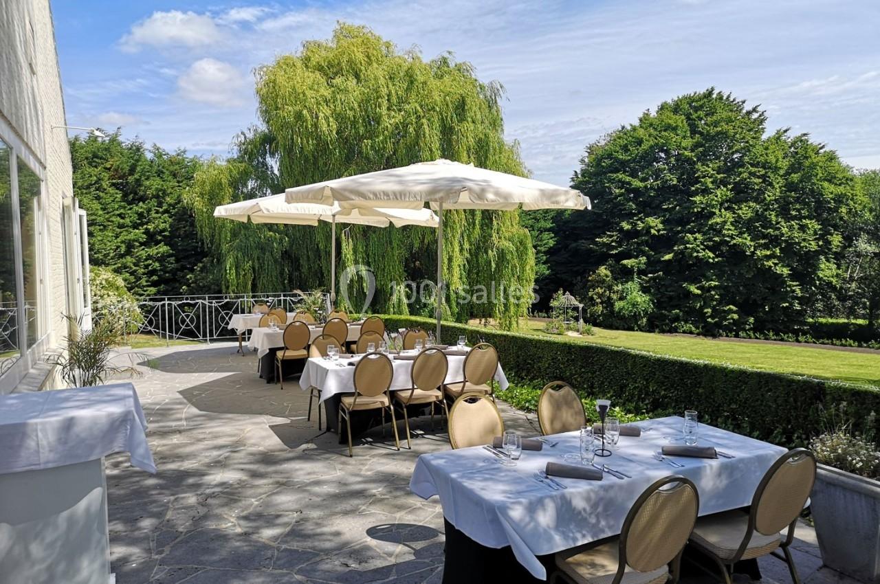 Terrasse d'un restaurant avec tables dressées, parasols blancs et vue sur un jardin arboré par une journée ensoleillée.