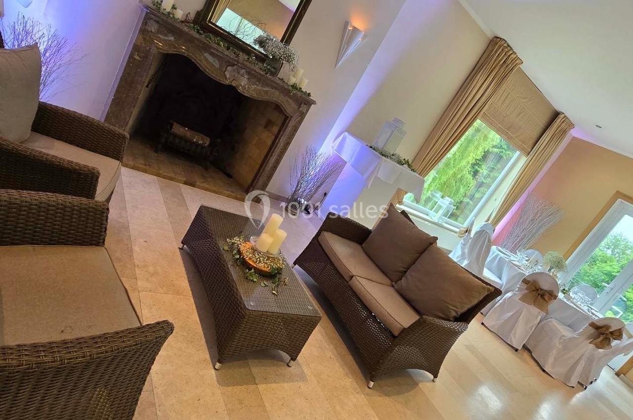 Salon lumineux avec fauteuils en rotin, cheminée décorative et vue sur une salle de réception avec grandes baies vitrées.