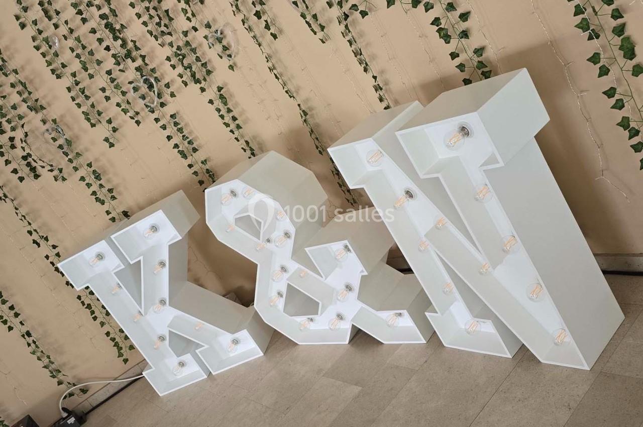 Lettres lumineuses ’K & N’ blanches posées au sol devant un mur décoré de guirlandes de lierre.