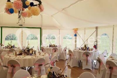 Deux balais déguisés en mariés avec robe et costume, posés devant une tente blanche et un cadre décoratif.