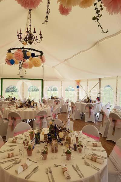 Salle de réception décorée avec des tables rondes dressées, des chaises ornées de rubans roses et des fleurs suspendues.