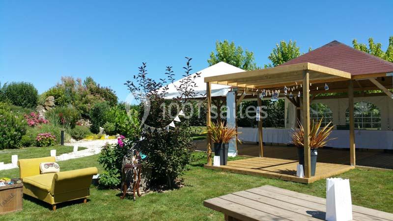 Espace extérieur avec tonnelle en bois, tente blanche, mobilier de jardin et végétation sous un ciel bleu.
