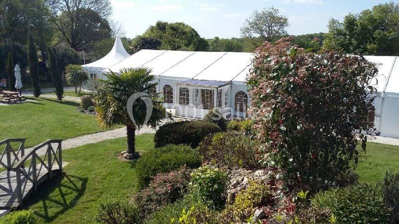 Vue d'un grand chapiteau blanc entouré de jardins verdoyants avec arbres, arbustes et un pont en bois.