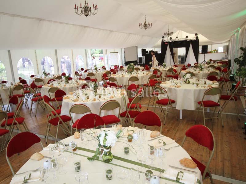 Salle de réception décorée avec des tables rondes dressées, chaises rouges et éclairage naturel sous une tente blanche.