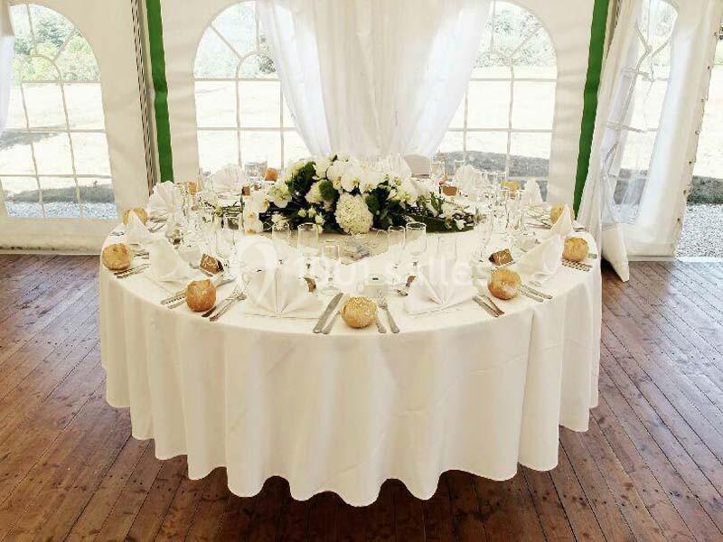 Table ronde élégamment dressée avec nappes blanches, vaisselle, fleurs et pains, dans une tente lumineuse.