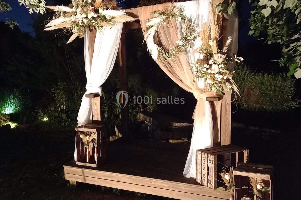 Arche de mariage en bois décorée de voilages blancs, fleurs et feuillages, éclairée dans un jardin en soirée.