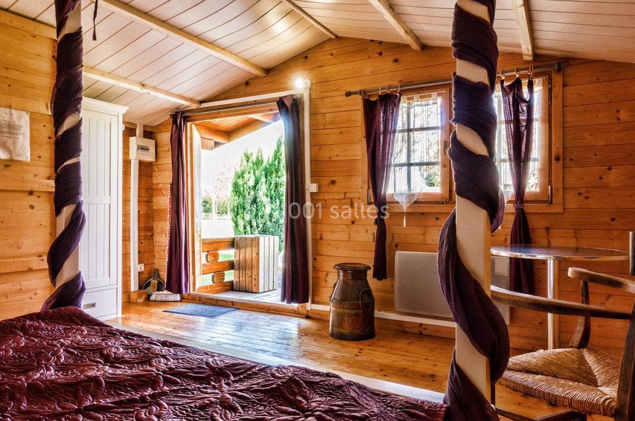 Intérieur d'une chambre en bois avec lit à baldaquin, rideaux violets, et vue sur une terrasse donnant sur un jardin.