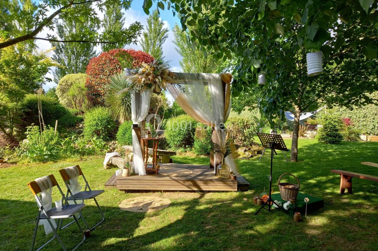 Arche de mariage décorée de voilages et de fleurs dans un jardin verdoyant, avec chaises et accessoires disposés autour.