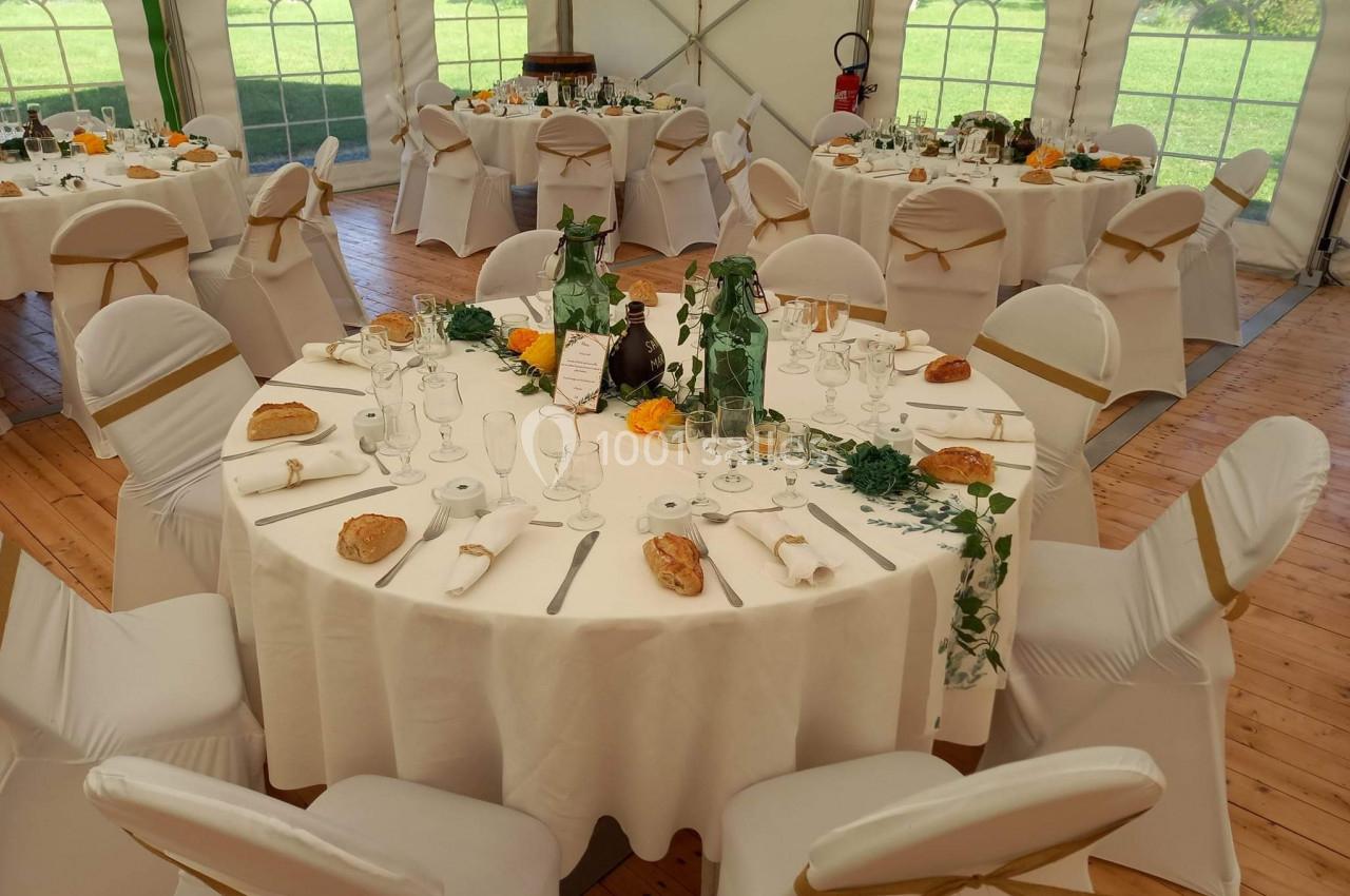 Tables rondes décorées pour un événement, avec nappes blanches, chaises houssées et éléments floraux dans une tente…