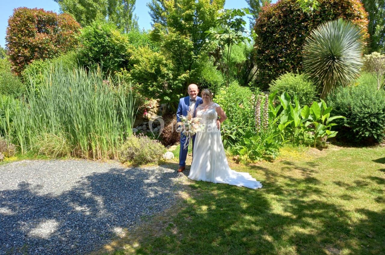 Un couple en tenue de mariage pose dans un jardin verdoyant sous un ciel ensoleillé.