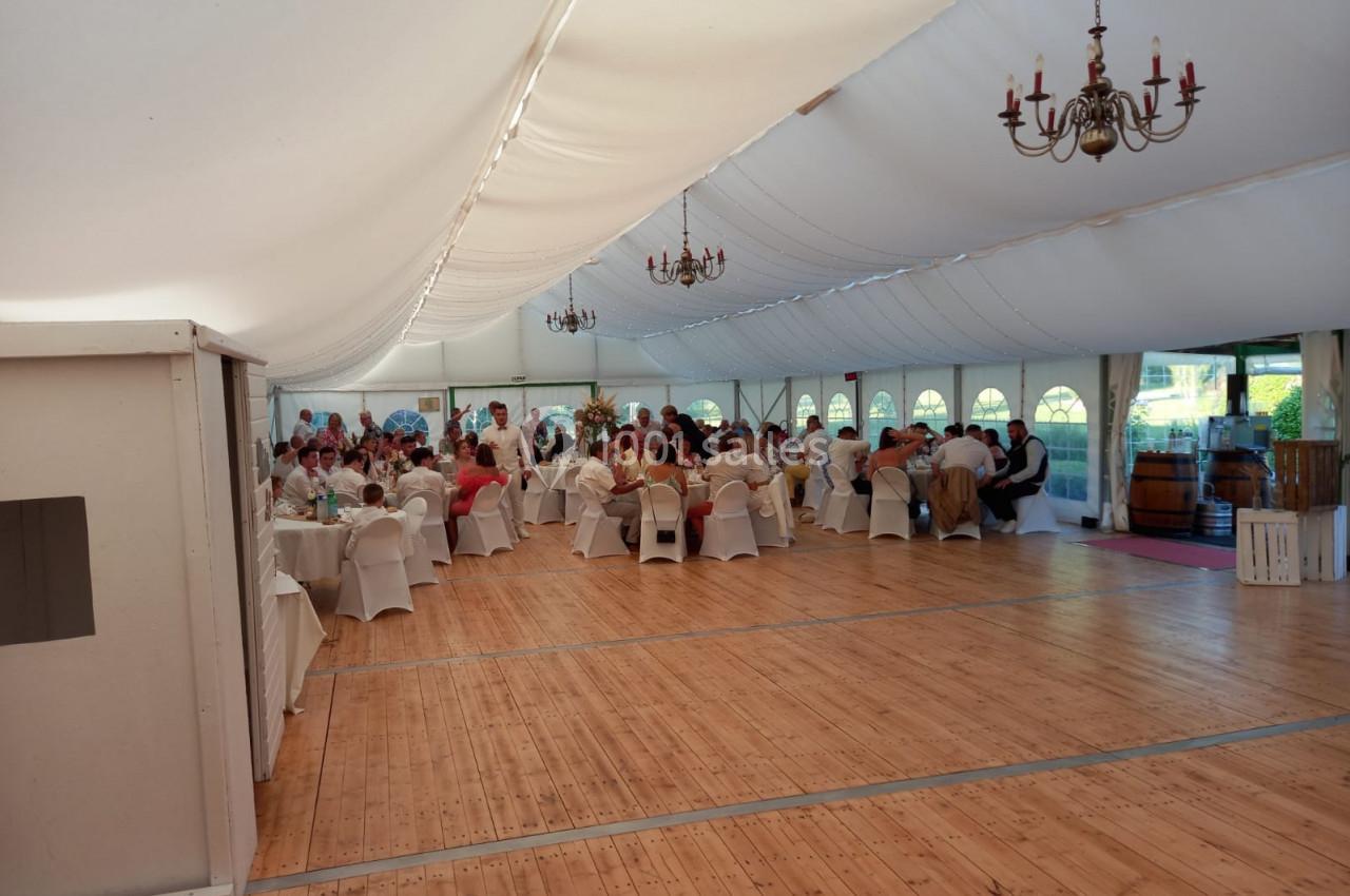 Salle de réception sous une tente blanche, avec des invités assis à des tables rondes et un parquet en bois.