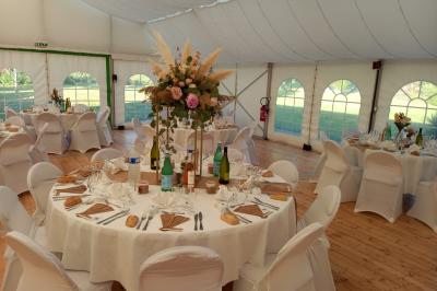 Deux balais déguisés en mariés avec robe et costume, posés devant une tente blanche et un cadre décoratif.